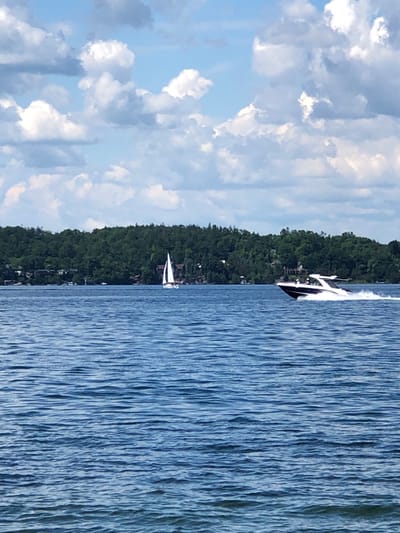 The image depicts a scenic view of Kempenfelt Bay in Barrie, Canada, on a partly cloudy day. The foreground is dominated by the calm, blue waters of the bay, with gentle ripples creating a textured surface. In the mid-ground, a white sailboat with its sail up is moving across the water. To the right of the sailboat, a sleek, white motorboat is speeding along, creating a visible wake. Along the distant shoreline, a dense, green treeline with scattered houses is visible. The sky above is a bright blue, dotted with fluffy white and gray clouds, suggesting fair weather.