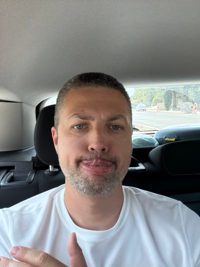 This media file is a selfie of a man in a car, taken from a slightly tilted angle. The man is wearing a white t-shirt and has short, dark hair with some grey stubble on his chin and cheeks. He is sticking his tongue out, with his eyes looking directly at the camera. The background shows the interior of a car, with a black headrest and grey upholstery visible. Outside the car window, greenery and some buildings can be seen, suggesting an outdoor setting. The lighting appears to be natural daylight, likely daytime. The man's expression seems playful or mischievous. No visible text is present in the image. The location context provided indicates Radanovići, Montenegro.
