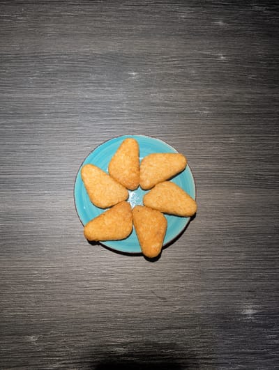 A top-down, full shot shows seven hash browns arranged in a circle on a turquoise plate with a brown rim. The plate is centered on a gray, wood-grain textured surface. The lighting is brighter on the left side of the image, casting a shadow on the right. There are no people visible. The location is not determinable from the image.