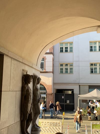 The image was taken in the city of Frankfurt am Main, Germany, at the coordinates 50.111 latitude and 8.683 longitude. The scene depicts a narrow alleyway, likely an urban passageway, with two stone statues of nude figures standing on a pedestal. The figures are facing forward, and the statues are partially lit by sunlight. The archway is made of concrete, with a curved ceiling. The scene is framed by the archway's opening, providing a view of the street beyond. On the opposite side of the street, a white building with multiple windows stands out. There are several pedestrians walking along the street, some of them appear to be interacting with a food stall or vendor, indicated by a white canopy tent. There is a bicycle leaning against the wall of the building on the left side of the image, with a few additional people standing behind it. The scene is framed by a large, concrete archway, which acts as a portal to the street beyond. The light is bright and sunny, indicating a time of day during the late morning or afternoon. The scene is mostly empty, except for the few people in the distance. The overall tone of the image is peaceful and serene.