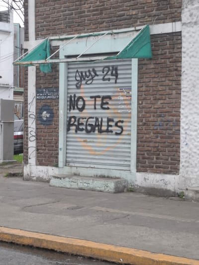 This image presents a street scene in Dock Sud, Argentina, centered on the lower portion of a weathered commercial building facade. The building features a reddish-brown brick wall interspersed with light-colored vertical and horizontal concrete or stucco elements. A closed, corrugated metal roller shutter, painted light grey, covers the main entrance. Black spray-painted graffiti on the shutter reads "Guys 24 NO TE REGALES," which translates to "Guys 24, DON'T GIVE YOURSELF AWAY." Faint, faded orange or yellow spray paint is visible beneath this text, and a small blue heart sticker is attached to the lower right side of the shutter. Above the shutter, a broken light-colored metal frame, presumably for an awning, has torn remnants of green fabric draped over it. To the left of the shutter, on the brick wall, there is a dark blue spray-painted circle containing a white cross-like or propeller-like symbol, alongside some indistinct, faded white text. A light-colored column further left is marked with black, vertically oriented, illegible graffiti.
The foreground consists of a grey concrete sidewalk, showing signs of wear, cracks, and minor patches of weeds growing from its edges and base of the wall. A yellow-painted concrete curb separates the sidewalk from the visible portion of the street, which appears to be wet or discolored. In the left background, part of a white building with an opening is visible, along with a portion of a silver or grey sedan parked on an unpaved area. Overhead wires are visible against the sky in the upper left. No individuals are present in the scene.