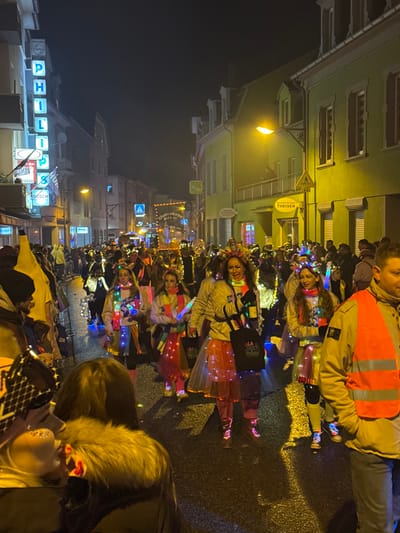 A nighttime scene depicts a festive parade in a city street, identified as Wasserbillig, Luxembourg. The wet street surface reflects artificial lights from buildings and participants. On the left, a multi-story building features a vertical neon "PHILIPS" sign and a "BOSCH" sign. On the right, a light green building has a balcony where an individual observes the parade, with a "THEISEN" sign visible below.

Numerous people participate in the procession, moving away from the viewer. Many wear brightly illuminated costumes, including light-up scarves, tutus, and headwear, indicating a carnival or similar event. Several women in the mid-ground are prominent, dressed in light-colored jackets, colorful light-up accessories, and translucent light-up skirts. One central woman holds a black bag with "LA FIESTA" branding and a white bottle-like object. Another participant wears a prominent light-up tiara. To the far left, a person in a large yellow banana costume is visible. In the background, the crowd is dense, with a banner partially visible bearing text that includes "Schendchehärz Witz el." In the foreground, a male figure on the far right wears a tan jacket and an orange reflective vest. Another person on the far left, seen from behind, wears a dark fur-lined hood. Streetlights illuminate the scene, casting shadows and reflections.