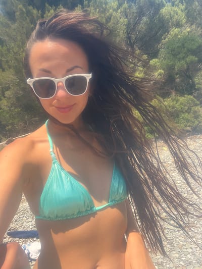 The media file shows a close-up, overhead shot of a woman wearing a turquoise bikini and white sunglasses. She has long, dark, wet hair that is blowing in the wind. Her skin is tanned and appears to be illuminated by the sun. The background consists of green foliage and a rocky, pebbly ground. The lighting suggests it is daytime, likely a sunny day. The scene appears to be outdoors, possibly at a beach or a scenic viewpoint, given the natural elements visible. The woman's face is partially visible, and she is smiling. The overall impression is one of a relaxed, possibly vacation setting. There is no visible text in the image.