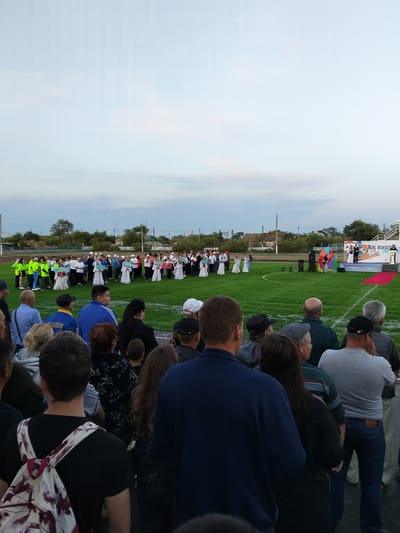 This is a medium shot, taken from a low angle, of an outdoor event, likely a festival or ceremony, taking place on a grassy field in Таман ауылдық округі, Kazakhstan. The sky is overcast with muted light, suggesting it is late afternoon or early evening. In the background, a small stage is set up with a banner that has Cyrillic text and Olympic-like rings. Two individuals in formal attire are standing on the stage, facing a crowd. To the left of the stage, a group of people in traditional white dresses and head coverings are holding rectangular signs, possibly representing different regions or organizations. Further to the left, a line of younger people in brightly colored athletic wear (green and yellow shirts) are also holding signs.
The foreground is dominated by a crowd of spectators, viewed from behind. Most are casually dressed in t-shirts, sweaters, and jeans. Some are looking towards the event on the field, while others appear to be conversing amongst themselves. The people in the crowd are diverse in age and gender. One person in the immediate foreground on the left is wearing a backpack with a floral pattern. The ground where the spectators are standing appears to be a darker surface, possibly a track or pathway around the field. The overall atmosphere seems to be one of communal gathering and celebration.