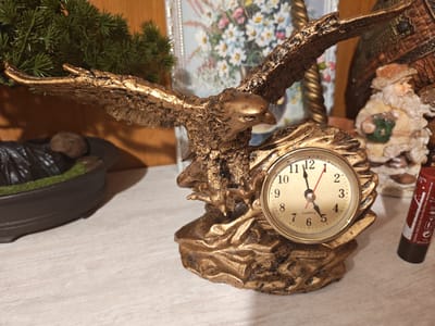 An ornate, metallic gold eagle statue with outstretched wings is positioned in the foreground, integrated with a small, round clock. The clock face has black numerals and hands, indicating a time close to 11:12. The statue rests on a textured, golden base. To the left of the eagle, a dark brown planter with a green moss-like substance and a grey stone is visible. In the background, a framed picture of flowers and a small, seated figurine resembling Santa Claus are present. A red tube, possibly lipstick or a similar cosmetic item with text in French, is positioned to the right of the clock. The scene is set on a light-colored, textured surface, possibly a table or shelf. The overall environment suggests an indoor decorative display. The location is stated to be in Чонкаўскі сельскі Савет, Belarus.