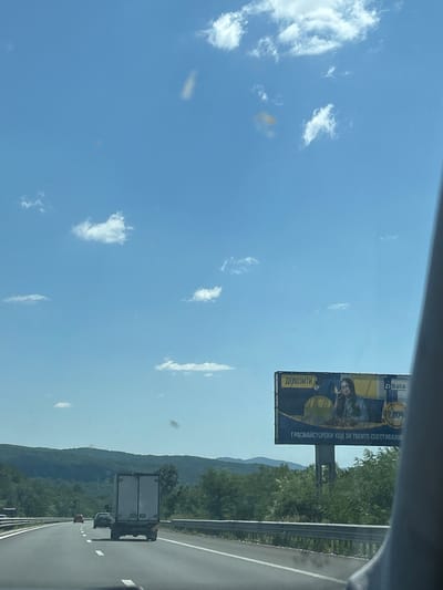 The image shows a highway in the city of Vakarel, Bulgaria, on a sunny day. The sky is clear blue with a few white clouds. On the side of the highway, there is a billboard advertising D Bank. The billboard features a woman and text in Bulgarian that translates to "Deposits" and "Great moves for your savings." There are several vehicles on the highway, including a white truck and some cars. In the background, there are green hills covered with trees. The overall scene depicts a typical day on a highway in a rural area.