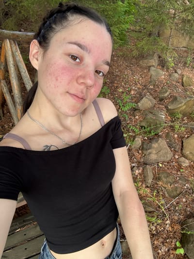 A young woman is taking a selfie outdoors. She is angled to the camera, with her face close to the lens. She has dark hair styled in a braid and is wearing a black off-the-shoulder top and denim shorts. A silver necklace is visible around her neck, and a small tattoo of a leaf design is on her neck. The woman's face has some redness and blemishes.
The background appears to be a natural setting with rocks, dry pine needles, and sparse green vegetation. There are also wooden planks visible, suggesting a rustic bench or platform. The lighting suggests it is daytime, possibly late afternoon, with dappled light filtering through the trees. The overall mood is casual and relaxed. There is no visible text in the image. The provided location context is Pozdyshki, Russia.