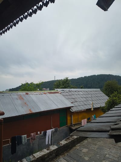 The image depicts a view in Dharamkot, India. A corrugated metal roof covers a multi-colored building with yellow, brown, and green sections. Clothes are hanging on a line attached to the building. Behind the building, there is a hillside covered in trees. In the background, a tall tower-like structure can be seen on the hillside. The sky is overcast. A portion of a roof with decorative trim is visible in the lower-left corner of the image. The perspective is from an elevated position looking down at the scene.