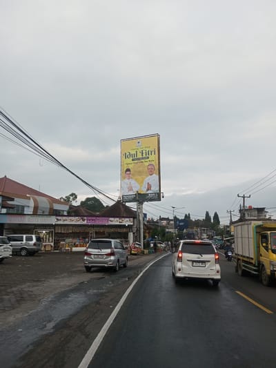 The image depicts a street scene in what appears to be a Southeast Asian town, possibly in Indonesia given the signage. A billboard advertising Eid al-Fitr is prominent, featuring portraits of two men in traditional attire. Roadside businesses are visible, including a shop called "Roti Unyil" and "Rahnita" which appear to sell local snacks and souvenirs. Several vehicles, including cars and a truck, are on the road. The sky is overcast, suggesting recent rain or an impending storm. Power lines crisscross the sky. The road surface appears wet. The license plate of the car directly in front of the camera reads "B 1473 NKX." The location in Taunggyi, Myanmar is not confirmed; the presence of Indonesian language and cultural references suggests otherwise.