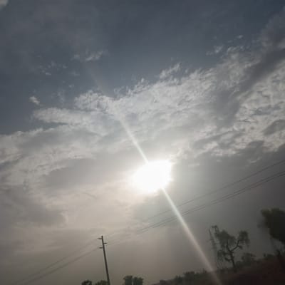The image shows a view toward the sky with the sun prominently positioned. Overcast, cloudy conditions prevail. In the upper right, trees and vegetation can be seen, alongside utility poles and power lines. The location is in the city of Chiromawa, Nigeria.