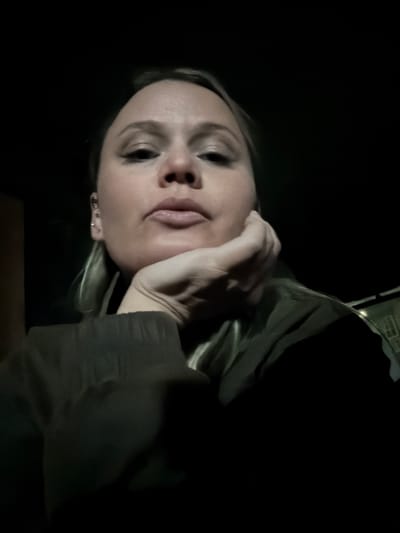 The image depicts an adult female in a dimly lit environment, purportedly in Dubnik, Russia. The subject, a fair-skinned individual with light-colored hair, occupies the majority of the frame, angled slightly from below, looking directly at the camera. Her right hand is observed with fingers curled, supporting her chin. She exhibits a neutral facial expression with slightly pursed lips. Multiple small metallic piercings are visible on her left ear. She wears a dark-colored garment, possibly a jacket or sweater. The background is almost entirely black, with a faint, indistinct structure or window outline barely perceptible in the lower right corner, suggesting artificial light or a distant reflective surface. The primary illumination appears to emanate from the front, highlighting the subject's face.