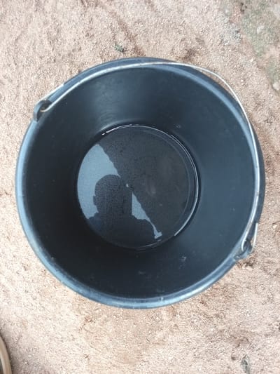 A black bucket with a metal handle sits on sandy ground. The bucket contains a small amount of water, reflecting the shadow of a person. The ground is a mixture of sand and small pebbles, with some sparse dried vegetation visible. The scene appears to be outdoors, possibly in a yard or unpaved area in Zinna, Nigeria.