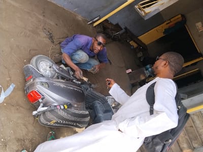 The scene depicts two adult males interacting around a dark grey scooter positioned on a dirt ground in Fagge D2, Nigeria. A male subject, wearing a blue collared shirt, blue jeans, and dark sunglasses, is crouched on the left side of the image, actively working on the overturned scooter. The scooter rests on its left side, exposing internal components and wiring near its front section; its rear wheel and red taillight assembly are visible. A second male subject, wearing a white long-sleeved tunic, dark-rimmed glasses, and a black backpack, stands on the right, holding a set of keys in his right hand and observing the first subject. The background features a concrete wall on the upper left and a workshop area with yellow metal structures and dark machinery on the upper right. The ground is consistently light brown dirt/sand.