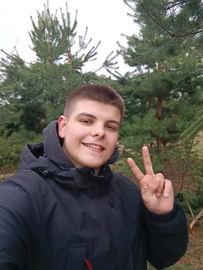 A young man is taking a selfie outdoors. He is smiling and making a peace sign with his right hand. He is wearing a dark-colored hooded jacket. Behind him are evergreen trees. Given the prompt context, the image was taken near Krasnyi Mayak, Russia. The environment appears to be a forest or wooded area. The weather looks overcast.