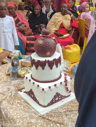 This image, captured in Unguwar Rimi, Nigeria, displays an outdoor social gathering with a prominent two-tiered cake as the central subject. The cake is positioned on a table covered with gold sequined fabric and a clear plastic sheet. It features white frosting with maroon-brown decorative details; the upper tier has a scalloped border, while the lower tier exhibits a beaded string and intricate scrollwork at its base. A dark reddish-brown ovoid cake topper with gold accents and maroon beads rests on the top tier.
To the left of the cake, a plastic-wrapped bottle with a heart emblem and partially visible "Mr." label is present, along with a woven basket containing small, individually wrapped items. In the background, behind the cake and to its right, two yellow plastic jerrycans are stacked, with several blue plastic water bottles visible below.
A group of individuals is seated in the background. From left to right: a young boy in a white traditional garment with blue embroidery looks slightly right; a woman wearing a salmon-colored headwrap and a striped dress holds a brown purse and displays a gold watch on her left wrist; another woman with a yellow headwrap is partially visible; a woman in a red headwrap and dark clothing holds a dark purse; a woman in a red dress with a cream head covering, also wearing a gold watch and holding a gold clutch, rests her chin on her left hand; and a woman in a pink headwrap and pink garment is seated in a yellow plastic chair. All individuals are dressed in traditional West African attire.