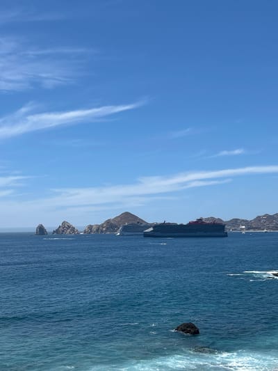 Captured from a vertical perspective, the image features a seascape likely located in Cabo San Lucas, Mexico. The scene is divided between a clear blue sky with high, wispy clouds and the deep blue expanse of the ocean. On the right side of the image, rocky cliffs and formations jut from the sea, defining the coastline. A large cruise ship is anchored off the coast, appearing between the cliffs and the open ocean. The water shows some signs of waves crashing against the shore, creating a foamy texture. The overall atmosphere is bright and sunny, typical of a coastal region.