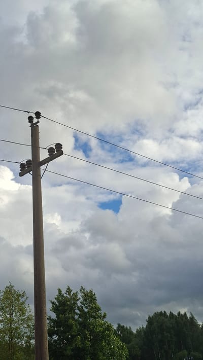 A concrete utility pole stands vertically on the left side of the image, supporting several horizontal power lines. The pole has two horizontal crossbars, each fitted with multiple ceramic insulators. Above the crossbars, the power lines extend towards the right. The sky is filled with a mix of dark, heavy clouds and lighter, scattered clouds, with patches of blue sky visible. Below the power lines and pole, a dense line of green trees forms the horizon. The scene appears to be in an outdoor, possibly rural or semi-urban, environment. The location is identified as the city of Smalyavichy, Belarus.