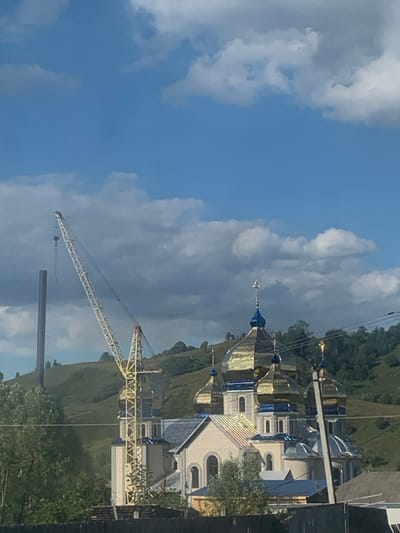The image displays the Transfiguration Church (Церква Преображення Господнього) in Lazeshchyna, Ukraine, situated within a mountainous rural landscape. The dominant subject is the church, characterized by multiple golden onion-shaped domes, several of which feature blue bases and are topped with Orthodox crosses. The church building itself is light-colored, with visible arched windows. To the immediate left of the church, a large yellow lattice-boom crane is positioned, with its boom extended upwards and a hook suspended from its end. Further left, a tall, slender dark grey or black industrial pipe or stack rises vertically. The background consists of rolling green hills covered with trees and vegetation. The sky is bright blue with scattered white and grey clouds. In the lower foreground, a dark horizontal barrier, likely a fence, is visible, partially obscured by green foliage. Overhead utility lines are present in the upper right quadrant of the frame. No individuals are visible. The presence of the crane indicates ongoing construction, renovation, or maintenance activities at the church.