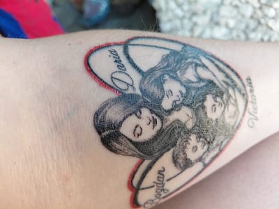 The image shows a close-up of a tattoo on a person's skin. The tattoo is in the shape of a heart and depicts four portraits of individuals, along with their names. The portraits are rendered in black ink with shading to give them depth. The outlines of the heart and some of the portraits are accentuated with red ink.
Text is visible within the tattoo: "Daria" is written in script on the upper left side, "Victoria" on the right side, and "Bogdan" on the lower side. The style of the portraits suggests a traditional or neo-traditional tattoo style. The skin appears to have some texture and slight redness around the tattoo, possibly indicating it is new or healing. The background is out of focus, but hints of clothing (pink and blue) and a textured surface (possibly concrete or stone) are present, suggesting an outdoor or casually lit setting. The overall impression is a personal and sentimental piece of body art.