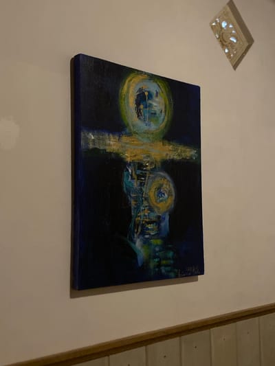 A vertical, dark blue canvas painting with abstract gold and blue markings hangs on a light beige wall. The painting features a dark background with swirling yellow, green, and blue hues that appear to form a circular shape at the top and a second, smaller circular shape in the center. A horizontal gold stripe is prominent across the middle of the painting. There is a small, decorative, rectangular object with cutouts mounted on the wall in the upper right corner. Below the painting, there is a wooden trim and then a section of wall paneling with vertical lines. The scene appears to be indoors, possibly in a residential or gallery setting. No people are visible in the image. The location in Buleleng, Indonesia is not discernible from the visual information provided.