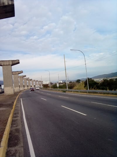 The image depicts a highway curving gently to the right. Along the left side of the road are several concrete pillars, apparently supporting an unfinished overpass or elevated roadway. A yellow curb separates the road from a strip of barren ground along the edge. White lines mark the lanes on the asphalt road. Two vehicles, a maroon car and a white van, are visible in the distance moving along the highway. On the right side of the road is a metal guardrail, separating the highway from an area of trees, shrubs, and utility poles. In the background, distant mountains are visible under a partly cloudy sky. The location is Parroquia Guatire, Venezuela.