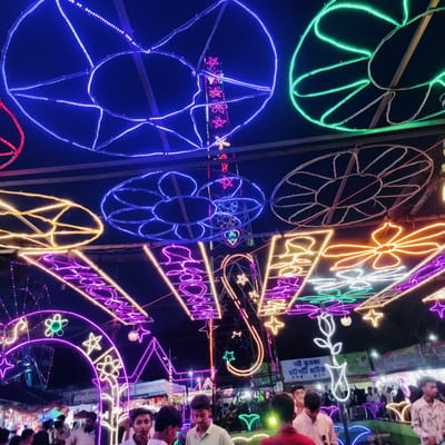 This nighttime photograph captures a vibrant fair or festival in Charfasson, Bangladesh, illuminated by an elaborate display of colorful LED lights. The scene is bustling with people, predominantly young men, gathered under a canopy of decorative lighting.

Overhead, numerous large light structures create a festive atmosphere. These include prominent blue and green circular patterns, some resembling abstract flowers or geometric designs, and others forming stars and hearts. Smaller, intricate light shapes in yellow, purple, red, and white are also visible, creating a dazzling array of illumination that stretches across the upper frame. A tall, central light structure features pink stars and other patterns, adding to the grandeur.

At ground level, a crowd of attendees can be seen moving amongst various stalls and vendors, typical of a lively market or fair. Visible signage in Bengali confirms the event's nature and location. Text like "বাণিজ্য মেলা" (Trade Fair), "জেলা প্রশাসক" (District Administrator), and "বাস মালিক সমি" (Bus Owners' Association) suggest organizers or participants. Food stalls are indicated by signs like "দই ফুসকা" (Doi Fuska) and "চটপটি হাউজ" (Chatpati House), offering popular local snacks. Another sign shows the brand "CLEMON" and "হাতসা এন্টারপ্রাইজ".

The overall mood is one of festive enjoyment and community gathering. The dark night sky provides a stark contrast to the brilliant, glowing lights, making them the focal point of the lively outdoor event.