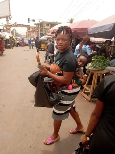 This image captures a bustling street scene in Umudioka, Nigeria, under daylight conditions. The central figure is an adult female walking towards the viewer's left, with a toddler secured to her back by a colorful fabric wrap. The woman has braided hair with black and orange strands, wears a black t-shirt, a grey and white striped knee-length skirt with a side slit, and pink beaded slide sandals. She holds a folded black umbrella with a wooden handle and a dark plastic bag in her left hand, and looks directly towards the camera with a neutral expression. The child, wearing a white top, looks over the woman's left shoulder directly at the camera.
To the right, a market stall features fresh green leafy vegetables displayed on a wooden stand, beneath large reddish-brown and dark blue market umbrellas. Numerous pedestrians are visible throughout the scene, indicating a busy commercial area. In the background, a paved road accommodates additional pedestrians, cars, motorcycles, and a yellow tricycle (keke napep). An overhead sign structure prominently displays the text "SOCIETY OF UMUDIOKA COURTESY CALL...", confirming the city location. A green traffic light is visible on a pole, and multi-story buildings line the distant street. Overhead power lines span across the upper portion of the frame.