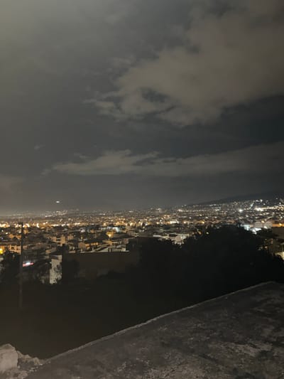 The image displays a panoramic nighttime view of Agia Paraskevi, Greece, from an elevated vantage point. The scene is dominated by a vast urban sprawl extending across the mid-ground and distant background, illuminated by countless warm-toned artificial lights emanating from buildings and street infrastructure. Hilly terrain, also dotted with lights, rises in the far right background. The sky above is dark and heavily overcast with visible, textured cloud formations. A solitary, bright light source is discernible in the far distance within the upper left quadrant of the sky. The immediate foreground features dark, indistinct foliage or trees on the left, partially obscuring the city, while a flat, rough, and textured concrete surface, likely a rooftop or balcony railing, occupies the lower right corner of the frame. No individuals, specific actions, or interactions are visible.