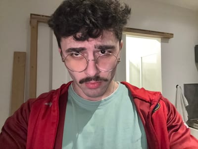 A man with a mustache and round glasses is facing the camera upside down. He is wearing a light blue t-shirt and a red jacket. A partially visible window and a wooden frame are in the background. A washing machine is slightly visible on the left. The image has a casual, possibly selfie style.