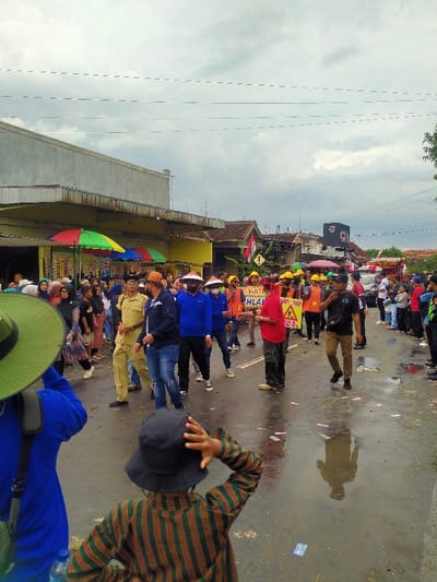 The image depicts a daytime outdoor scene on a wet asphalt road in Mojogedang, Indonesia, under an overcast sky. A procession of individuals is moving along the street from right to left.
In the immediate foreground, positioned with their backs to the viewer, an adult in a blue shirt wearing a wide-brimmed green hat stands on the left. To their right, a child in a brown and green striped long-sleeve shirt and a black bucket hat stands with their right hand raised to their head, observing the scene ahead.
The main subject is a group of approximately twenty adults in the midground, walking on the wet road. Several individuals within this group wear yellow hard hats and bright orange or red safety vests. One man is dressed in a full khaki uniform. Multiple people are jointly carrying a large yellow banner with black text, partially visible as "HATI-HATI" (Be careful), and additional obscured Indonesian words. Another orange banner is partially visible behind it. On the right side of the street, a line of spectators observes the procession. The street itself is visibly wet and reflective, with small scattered pieces of light-colored debris.
To the left, along the street, a long building with a light-colored facade features a covered walkway. Below the walkway, several vendor stalls are indicated by bright red, green, and blue umbrellas. Further down the street, on both sides, various single and multi-story structures are visible. A yellow triangular warning sign with an exclamation mark and a black rectangular sign are visible on the right side of the street. A white vehicle is partially visible parked further down the road on the right. Overhead, multiple utility lines stretch across the sky.
