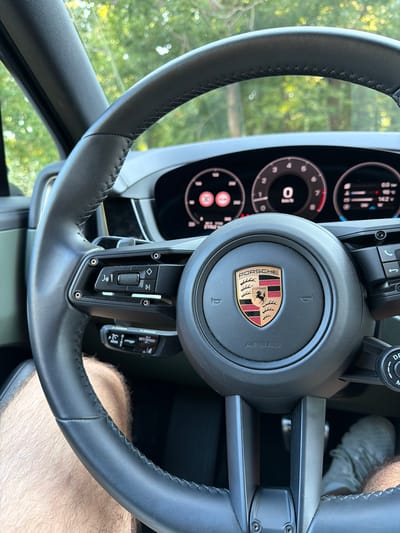 The image shows a close-up interior view of a Porsche steering wheel and dashboard. The steering wheel is black leather with stitching and features the Porsche logo in the center. Buttons and controls are visible on the steering wheel, including volume and cruise control settings. The dashboard displays a digital instrument cluster with a speedometer showing 0 km/h, a tachometer, and other vehicle information. The exterior scene visible through the windshield consists of blurred green foliage, suggesting an outdoor environment. The image is taken from the driver's perspective. The location in Lozenets, Bulgaria, is not discernible from the visual information provided.