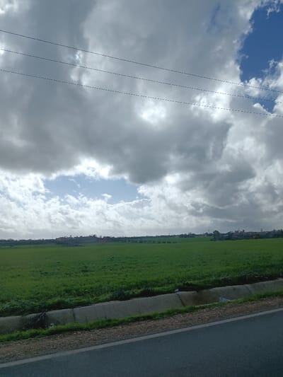 The image depicts an expansive rural and urban landscape in Sidi Slimane Moul Al Kifane, Morocco, under a heavily clouded sky. The foreground features a dark grey paved road surface on the bottom right, adjacent to which is a concrete-lined drainage ditch with sparse green vegetation and some visible debris. Beyond the ditch, a vast, flat field of vibrant green crops, possibly agricultural farmland, extends across the mid-ground. On the distant horizon, a scattered outline of structures, including residential buildings and possibly a minaret, indicates the presence of a town or city, partially obscured by distance and atmospheric haze. Several trees are also visible near the urban outline. The sky above is dominated by numerous large, grey, and white cumulus clouds, with prominent patches of clear blue sky visible to the upper right and through a significant opening in the cloud cover in the central-left portion, where sunlight appears to break through. Three parallel dark utility wires traverse horizontally across the upper third of the frame. No individuals, specific actions, or interactions are observable within the scene.