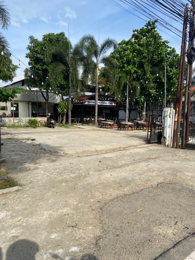The image shows a view of a street in Purwakarta, Indonesia, located at the coordinates -6.552 latitude, 107.437 longitude. The scene is an empty parking lot in front of a small restaurant. The street in front of the parking lot is paved with asphalt. On the right side, the street is lined with a fence, a large metal gate, and a tall metal pole, with a few power lines hanging from it. The parking lot is made of gravel, and the parking lot is bordered by trees and foliage, including palm trees and leafy green trees, and several electrical wires are strung between them. There is a small, brown motorcycle parked in the parking lot. The restaurant building is visible in the background, with a sign that reads "WARKOP ROPANG PWK". There are several tables and chairs in front of the restaurant. The restaurant is surrounded by foliage and appears to be closed, with the lights turned off.