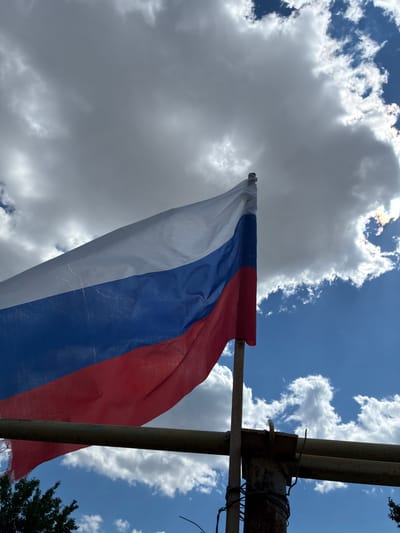 The image shows a flag with three horizontal stripes: white on top, blue in the middle, and red on the bottom. The flag is attached to a metal pole and appears to be waving in the wind. The background is a sky with a mix of white, fluffy clouds and patches of bright blue. Sunlight is breaking through some of the clouds, creating bright spots and rays. The flag is positioned in the lower right portion of the frame, and its colors are vibrant against the sky. Some of the clouds are quite dense and gray, while others are thinner and more wispy. The overall impression is of a bright and possibly windy day. The setting appears to be outdoors, with the flag likely displayed in a public space or on a property. There are no people visible in the image. A sliver of dark green foliage, possibly a tree, is visible in the far upper right corner. The flag is somewhat frayed at the bottom edge. The scene suggests a daytime setting, given the brightness of the sun and the blue sky. The presence of the flag might indicate a national celebration or simply be a patriotic display.
