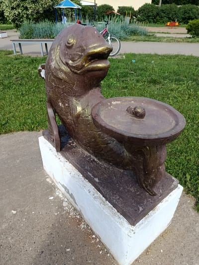 A weathered, bronze-colored statue of a fish with gold accents sits on a concrete pedestal in a grassy area. The fish is depicted in a seated position, holding a shallow dish on its lap with two small, round objects within it. The statue is in a park-like setting, with a paved path, benches, and playground equipment visible in the background. A bicycle is also parked nearby. The environment suggests an outdoor public space. The statue appears to be part of a larger decorative element or children's play area.