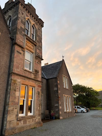 This image captures a multi-structure building complex in Sconser, United Kingdom, at what appears to be sunset. The scene is dominated by a two-part building and a gravel forecourt, with natural landscape in the background.

The primary structure on the left is a stone-built tower-like section, featuring irregularly cut, rusticated stone blocks in shades of grey, brown, and reddish-brown. The tower has multiple levels of white-framed windows, with the lower windows prominently reflecting the warm, orange-yellow light of the sky. The top of the tower exhibits a crenellated parapet design. A darker, rendered section of the building is partially visible to the immediate left of the stone tower.

Adjacent and to the right of the tower is a separate, smaller gabled building with a dark, textured, rendered exterior and a dark, pitched slate roof. A decorative metal finial or weather vane is positioned at the apex of its gable. This building also features white-framed windows. Between the two buildings, a small patio area includes a wooden chair and several potted plants.

The ground in the foreground and midground is covered with light grey gravel, forming a parking area. Three vehicles are parked on the gravel: a white minivan in the midground, a dark-colored car partially obscured behind it, and a third dark vehicle on the far left.

The sky occupies the upper portion of the frame, transitioning from a light blue-grey at the top to soft pinks, oranges, and yellows towards the horizon, indicative of a sunset. Wispy clouds are distributed across the warm-toned sky. In the background, beyond the buildings and vehicles, a large, mature tree with dense green foliage is visible. Distant green hills or elevated terrain are discernible on the horizon to the right. A small, round red object (possibly a buoy or similar marker) is partially visible hanging near the tree line. A small white sign displaying "VACANCIES" in dark text is affixed to the mullion of a lower window on the stone tower.