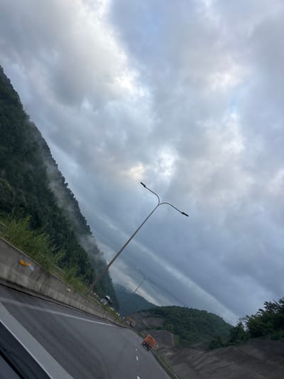 The image captures a scenic view of a highway in Tsakva, Georgia, on a cloudy day. The highway runs alongside a lush, green mountain that is partially shrouded in mist, creating a dramatic and atmospheric effect. The road is wet, suggesting recent rain or high humidity. Several vehicles, including trucks and cars, are visible on the highway, indicating ongoing traffic. A street lamp with two lights stands tall on the right side of the road, its dark silhouette contrasting with the bright sky. The sky itself is filled with heavy, grey clouds, with patches of lighter sky peeking through, hinting at the possibility of the sun breaking through. The overall impression is one of natural beauty and a sense of journey, with the mountain range and the cloudy sky dominating the landscape.
