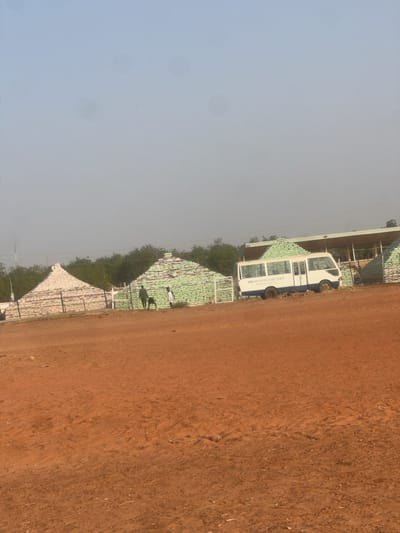 The scene, located in Birnin Kebbi, Nigeria, presents an outdoor landscape under a clear, pale blue sky. The foreground consists of reddish-brown, dry earth. In the midground, multiple conical or pyramidal structures are evident, constructed from stacked bags. The leftmost prominent structure is composed of light-colored bags, while an adjacent structure to its right is made of light green bags. A low, rudimentary fence, formed by vertical posts and horizontal elements, encircles these structures. A white minibus, featuring a blue stripe and the partially visible text "MINISTRY FOR SPORTS" on its side, is parked beside the green-bagged structure. Behind the structures, a line of green trees is visible, alongside a simple, open-sided shelter. Two human figures are present near the green-bagged structure: one in dark clothing, the other in lighter attire. A small, dark, four-legged animal stands between the two individuals.