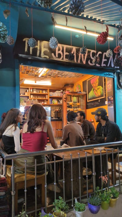 The image shows the exterior of "The Beanscene" coffee shop in Dharamkot, India. People are visible inside the shop and standing outside on a staircase with metal railings. Potted plants decorate the stairs. The shop's sign hangs vertically and is adorned with pine cones. Inside, shelves are stocked with various items, and artwork is displayed on the walls. The overall lighting suggests an evening or nighttime scene.