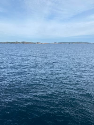 This is an aerial, landscape-oriented video clip shot at Stavern, Norway. The scene is dominated by the expansive blue sea, stretching across the majority of the frame. Gentle ripples and waves create a textured surface on the water, which appears to be a deep, rich blue. To the left of the frame, a sliver of coastline is visible, characterized by rugged terrain and green vegetation. Beyond the coastline, a clear blue sky occupies the upper portion of the image, with faint, wispy clouds scattered across it. The overall impression is one of a serene and vast natural landscape, likely captured on a clear day. The video is oriented vertically, suggesting it was filmed while looking down from a higher vantage point such as a drone or helicopter. There is no discernible activity or text within the frame.