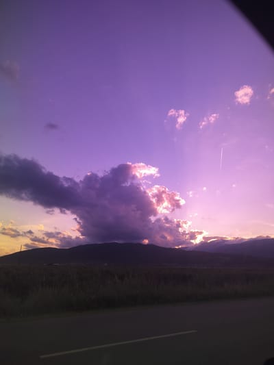 This is a photograph taken in Manolovo, Bulgaria, capturing a sunset over a rural landscape. The sky is dominated by shades of purple and pink, with fluffy clouds illuminated by the setting sun. A dark silhouette of mountains runs across the horizon, separating the sky from a field of tall grass and vegetation. A paved road with a white line runs diagonally across the bottom of the image, suggesting the vantage point is from a vehicle. A contrail from an airplane is visible in the upper right portion of the sky.