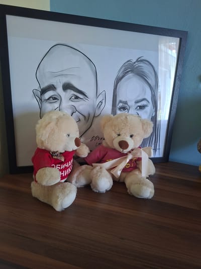 Two cream-colored teddy bears are positioned on a wooden surface in front of a framed caricature. The teddy bear on the left wears a red shirt with white Cyrillic lettering that appears to read "ОБИЧАМ НИКИ". The teddy bear on the right wears a pink shirt adorned with a pale pink ribbon tied in a bow. The caricature behind them depicts two faces, one larger and closer, with prominent features, and another partially visible. The background suggests an indoor setting, with a teal-colored wall visible to the right. The overall scene is brightly lit, with the focus on the teddy bears and the caricature. This image appears to be taken inside a home in Aksakovo, Bulgaria, possibly on a shelf or table.