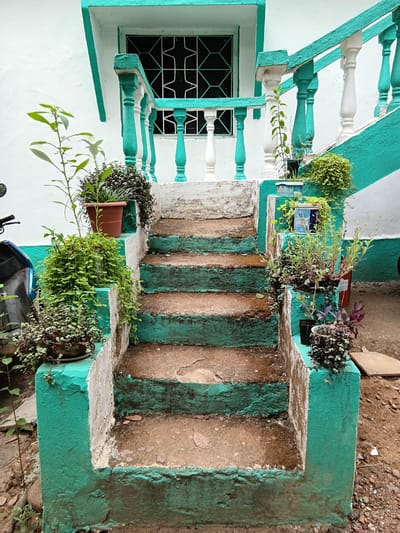 The image depicts a set of outdoor stairs leading to a building, possibly a house. The stairs are constructed of concrete and painted with alternating stripes of turquoise and a weathered, brownish-tan color. The paint shows signs of age and wear. Several potted plants and other greenery are positioned along the sides of the stairs and on the adjacent walls. The wall of the building is white with turquoise trim around a window that has a black, geometric-patterned security grille. The scene is located in Badem, India. The ground adjacent to the stairs is dirt and pebbles. A partial view of a vehicle, possibly a scooter or motorcycle, is visible at the bottom of the image. The overall environment is one of a residential area with older buildings.