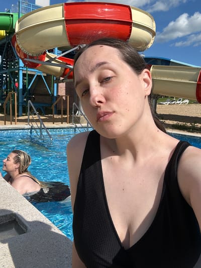 A woman is taking a selfie in a swimming pool at a water park. She is wearing a black swimsuit and has brown hair. In the background, there are water slides and lounge chairs. Another woman with blonde hair is in the pool. The sky is blue with some clouds. The location is the city of Yubileyny, Russia.