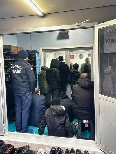 The image shows a group of people gathered in what appears to be a small prayer room or community center. The lighting is artificial and somewhat dim. In the foreground, a person wearing a dark blue uniform with "KOCAELI İTFAİYE" and a Turkish flag emblem is standing, facing away from the camera. They are wearing a dark blue cap and dark blue pants. To the left, there are shelves filled with shoes, indicating a place where shoes are removed before entering.
The majority of the individuals in the image are seen from behind, dressed in casual winter clothing, suggesting a cooler climate. Some are standing, while others are sitting or kneeling on a blue carpet that covers the floor inside the room. The room itself is modest, with a doorway leading into another area where more people are visible.
On the right side of the image, a sign is partially visible with the word "KURU" in large letters. There are also some reflections in the glass door. The overall impression is of a gathering for prayer or a community event. The presence of shoes on shelves outside the room and the carpeted floor inside suggest a custom of removing shoes before entering a sacred or communal space. There is no specific visual information in the image that directly identifies the location as Harmanli, Bulgaria.
