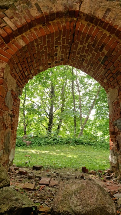 The image depicts an arched opening in a ruined brick structure, looking out onto a lush, green outdoor scene. The foreground is dominated by weathered red bricks, forming the archway, with some rough stones and rubble visible along the edges of the opening. The bricks show signs of age and decay, with visible mortar and some cracks.

Through the arch, a bright, sunlit area of green grass and dense foliage is visible. Several trees with full, green canopies are prominent, their branches reaching upwards and outwards. The sunlight filters through the leaves, creating dappled patterns on the ground. The scene suggests a warm, possibly summer day. The overall impression is one of nature reclaiming an old, man-made structure. There are no people or discernible text visible in the image. The location is contextually identified as Embūte, Latvia.
