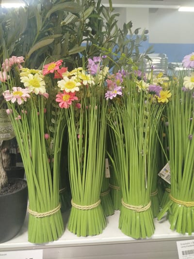 This is a photograph of an assortment of artificial flowers and greenery displayed on a shelf. The subjects are primarily artificial plants, including bunches of long, green, grass-like stems tied with twine, interspersed with various colorful artificial flowers in shades of pink, yellow, and purple. There are also some larger, leafy green branches in the background, possibly part of an artificial olive tree.

The setting appears to be a retail environment, likely a store display, indicated by the shelf and the presence of product labels. The lighting is bright and even, suggesting indoor illumination, possibly from overhead fluorescent lights. There are no people visible in the image, and no specific activity or event is taking place; it is a still-life shot of merchandise.

Notable details include the texture and material of the artificial plants. The green stems have a somewhat plastic or rubbery appearance, and the flowers are vibrantly colored. The twine used to bundle the stems is a light, natural color and has a woven texture. In the background, out-of-focus elements include what appear to be more shelves or displays, and a blue wall.

Visible text includes a label on the left side with "HOME ACCENTS" and "erial Olive" partially visible. On the right side, a label is present with German text, possibly related to product information. A price tag is visible at the bottom of the image with "0,27". The context provided indicates the location is Offenbach am Main, Germany. The overall impression is a colorful and artificial floral arrangement presented for sale.
