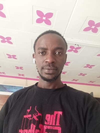 The image shows a close-up selfie of a Black man with a short beard wearing a black t-shirt with a red design. The man is looking directly at the camera. He is wearing a white earbud in his left ear. The ceiling is white with pink four-petaled flower designs. The background of the photo suggests an interior room, possibly a residence in the city of Ganja, Azerbaijan, based on the provided coordinates.  The image appears to be a casual selfie captured by the individual in the image.