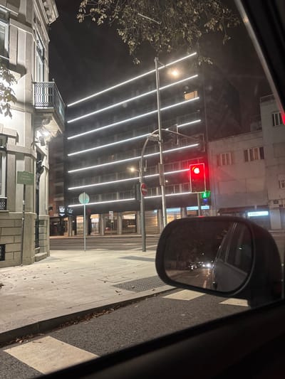 The image depicts a nighttime street scene from inside a stationary vehicle at the intersection of Rua Alegria Junqueiro and Rua de Dom Pedro V in Lordelo do Ouro e Massarelos, Porto, Portugal.
To the left, a multi-story building with a light-colored stone facade and an ornate black wrought-iron balcony is visible. A white street sign on its corner clearly reads "RUA ALEGRIA JUNQUEIRO." Directly ahead, a modern, dark-colored multi-story building dominates the background, characterized by multiple parallel horizontal white LED light strips illuminating its facade.
A tall street light pole stands centrally, supporting a vehicle traffic light displaying a solid red signal and a pedestrian traffic light displaying a green walking figure. Below these, a circular red 'no entry' sign is visible. The asphalt road features white pedestrian crossing stripes. The concrete sidewalk to the left is partially covered with scattered brown leaves.
The foreground includes the interior bottom frame of a vehicle window and the passenger-side exterior mirror. The mirror's reflection shows a dark street behind the camera's position, with the headlights of several distant vehicles visible. The overall scene is devoid of people.