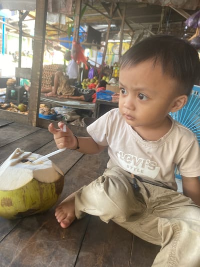 A young child, appearing to be of Indonesian descent, is seated on a weathered wooden surface, possibly a pier or market stall, in what appears to be an outdoor setting within the city of Ta', Indonesia. The child is holding a white straw inserted into a halved green coconut, which rests on the wooden surface to their left. The child is wearing a light-colored t-shirt with a graphic that reads "Levi's Style" and khaki-colored pants. The child's right hand is holding the straw, and their left hand is near their mouth. The background suggests a bustling market environment, with structures made of wood and corrugated metal overhead, and various goods such as fruits and fabrics visible in the distance. Other individuals are present in the background, some appearing to be resting or working. A blue fan is visible to the right of the child. The lighting indicates it is daytime, with sunlight filtering through the structures.